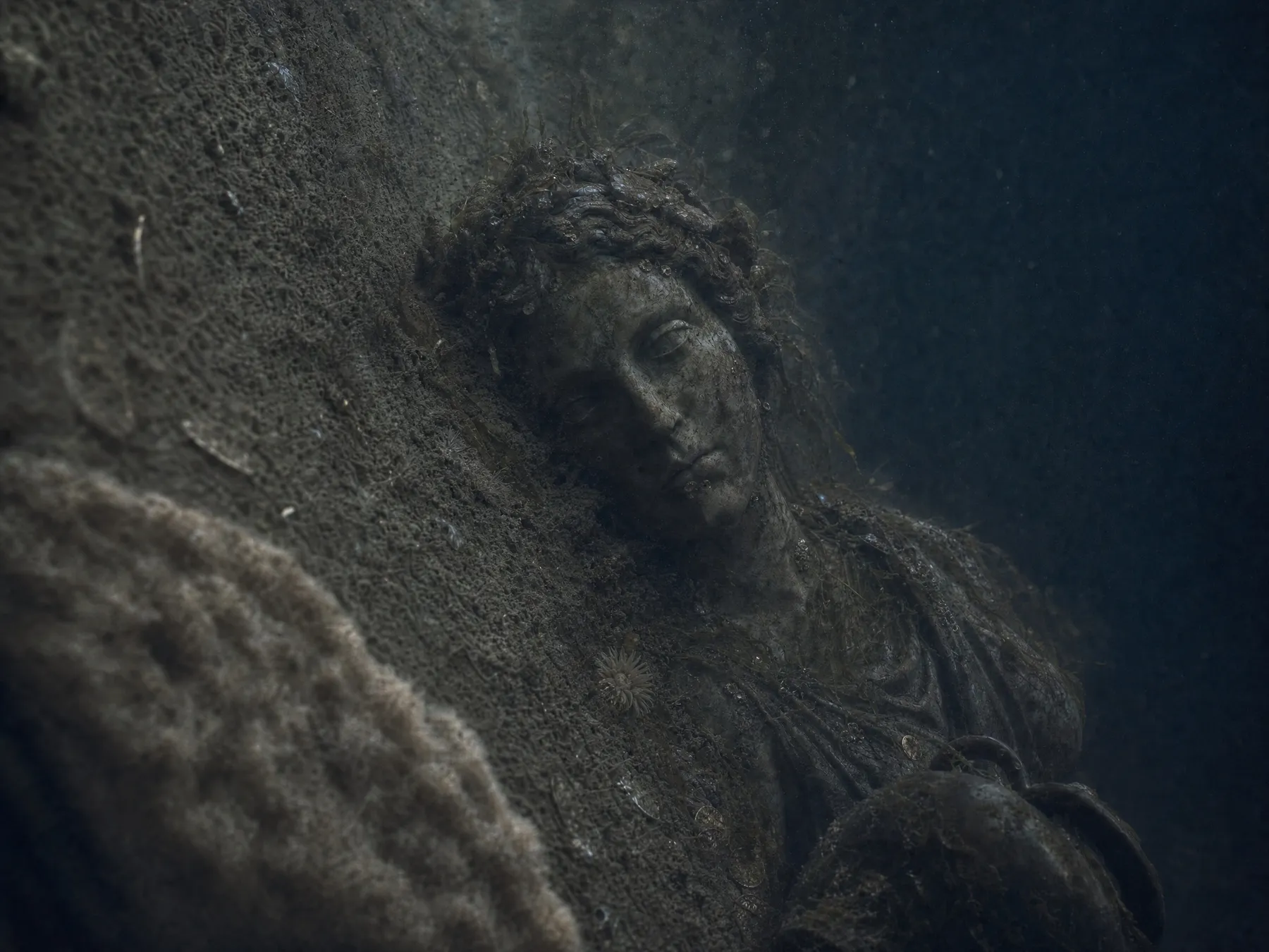 Persephone lies on her side on a dark seabed, weathered and covered in growth